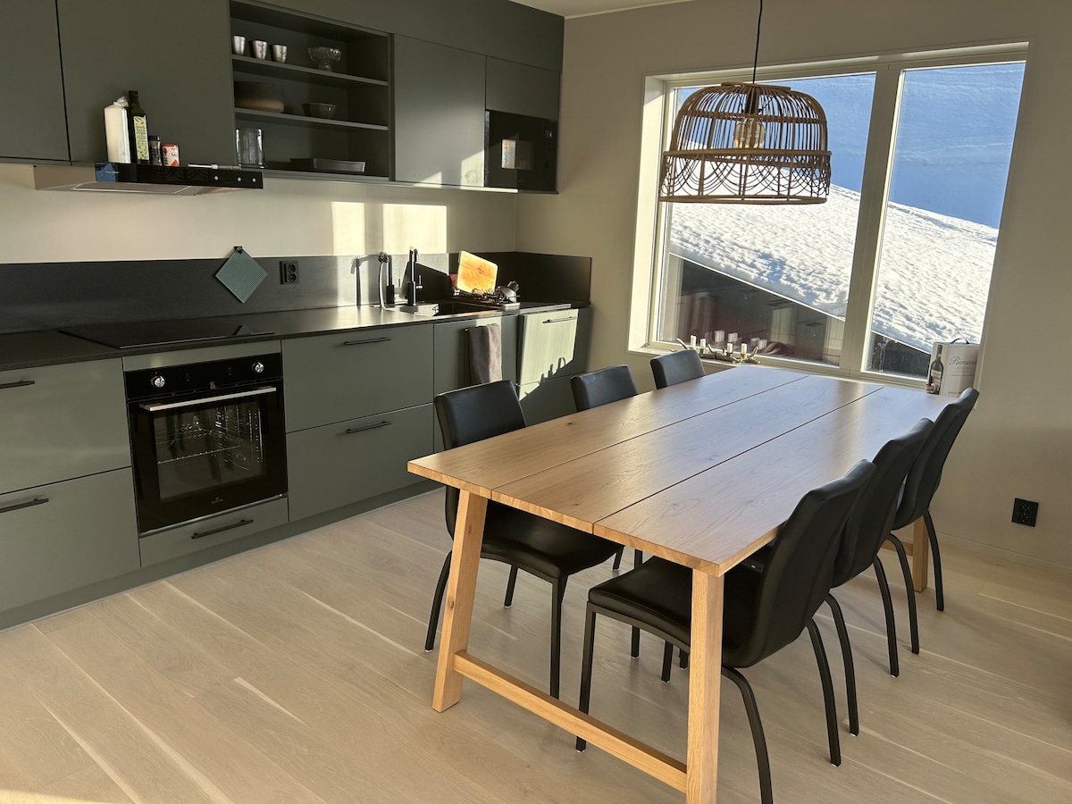 A modern kitchen is equipped with sleek cabinetry and appliances, including an oven and a microwave. A spacious wooden dining table with six black chairs is positioned by a large window, allowing natural light to illuminate the room. Snow-capped scenery can be seen outside.