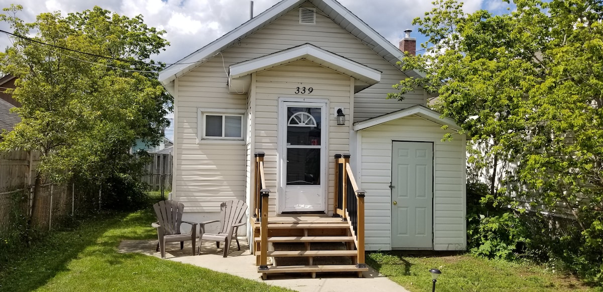 The exterior of the house is presented, featuring a welcoming front porch with steps leading to the main entrance. A small storage shed is visible to the right, and two outdoor chairs are positioned on the grassy area, surrounded by greenery.