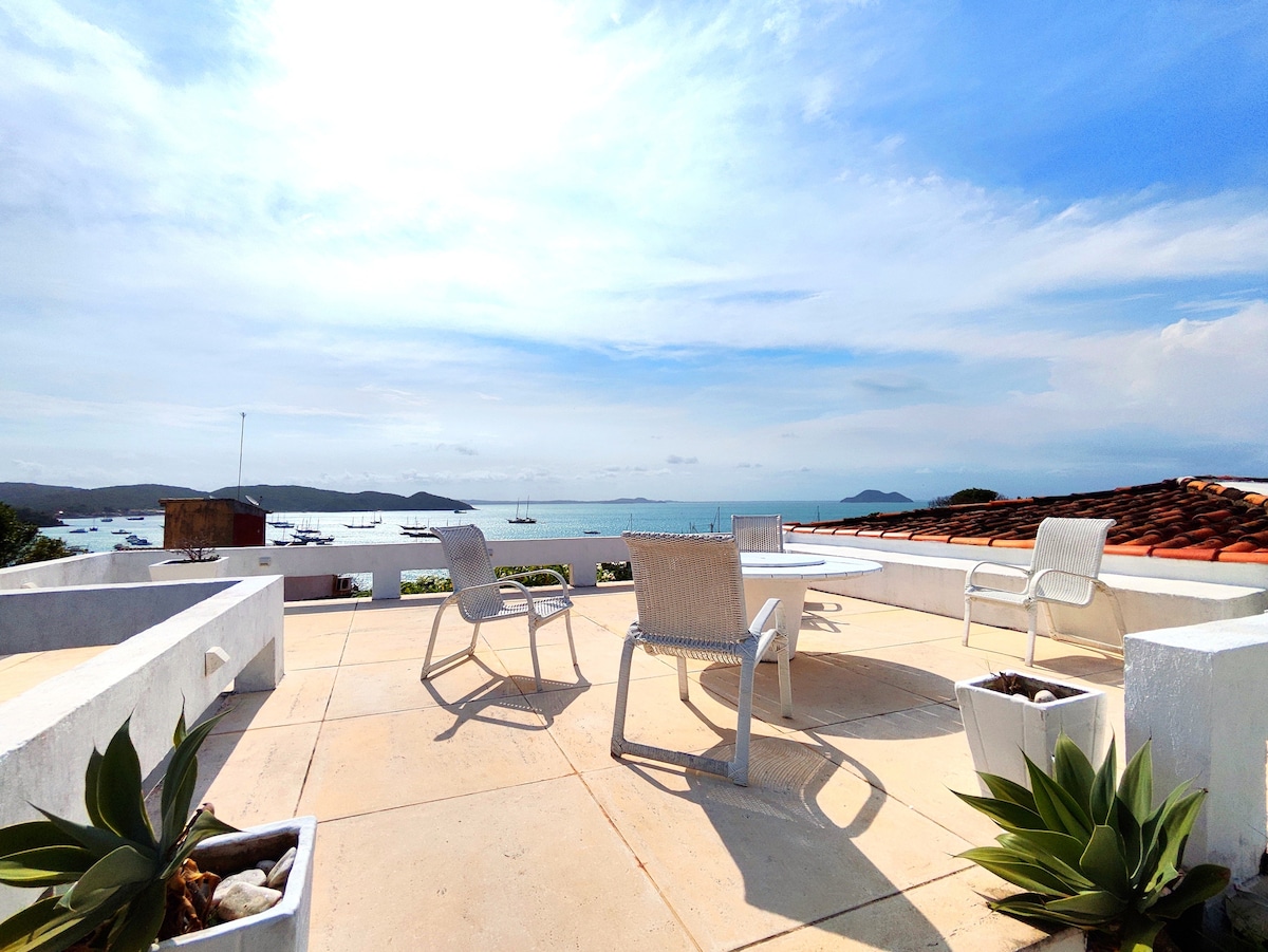 A spacious terrace offers stunning views of the ocean, with white stone flooring and bright blue skies. Four simple white chairs surround a round table, complemented by potted plants, creating a serene space for relaxation.