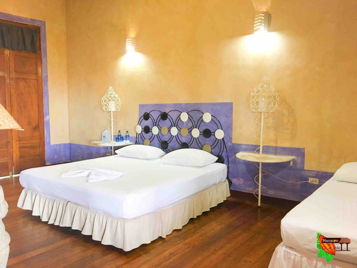 A well-lit sleeping area features a large bed with a white ruffled bed skirt and two bedside tables, each designed with a unique base. A second smaller bed is positioned nearby. Warm tones of the wall complement the wooden floor, creating a cozy environment.