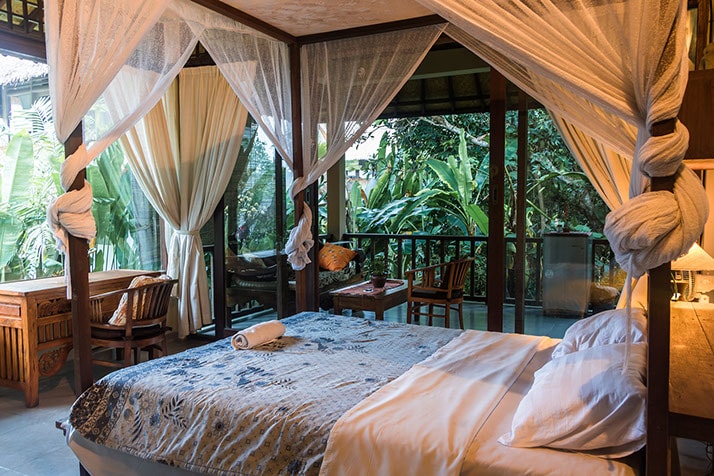 A cozy sleeping area is presented with a spacious bed draped in light fabric, surrounded by greenery visible through large glass doors. A writing desk and wooden chairs are positioned nearby, enhancing the open and inviting ambiance.