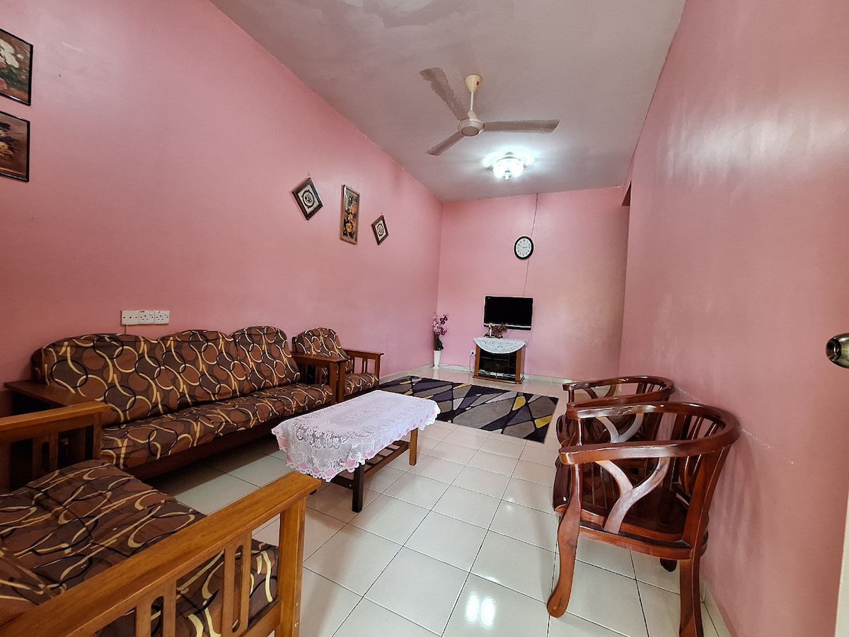 A living area is presented with light pink walls, featuring a patterned sofa set complemented by two wooden chairs. A small white table, draped with a lace cover, is positioned in the center. A television is mounted on the wall, providing entertainment options for guests.