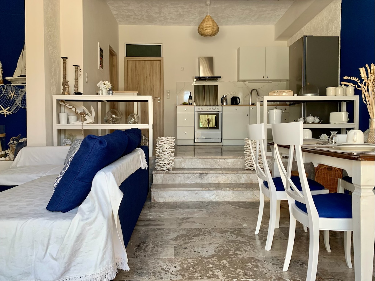 The open-concept living space features a stylish kitchen with stainless steel appliances and white cabinetry. A dining table with white chairs is visible, alongside a comfortable seating area adorned with blue accents. Natural light enhances the inviting ambiance throughout the room.