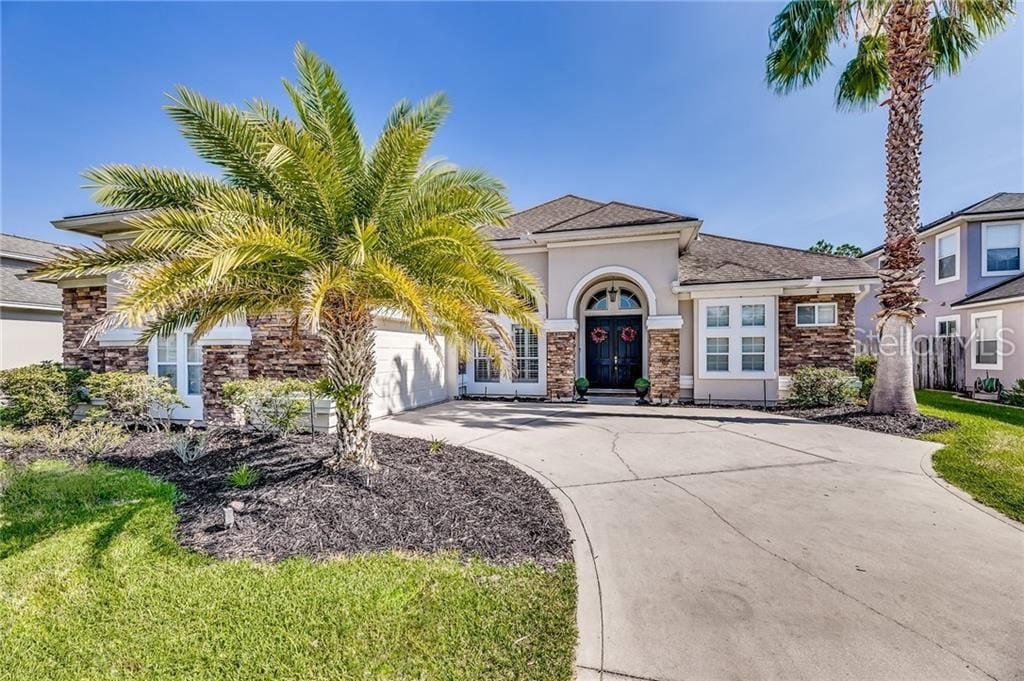 The exterior of a single-story house is shown, featuring a curved driveway lined with dark mulch and manicured grass. Palm trees are positioned near the entrance, which is accentuated by a decorative doorway and two arched windows on either side.