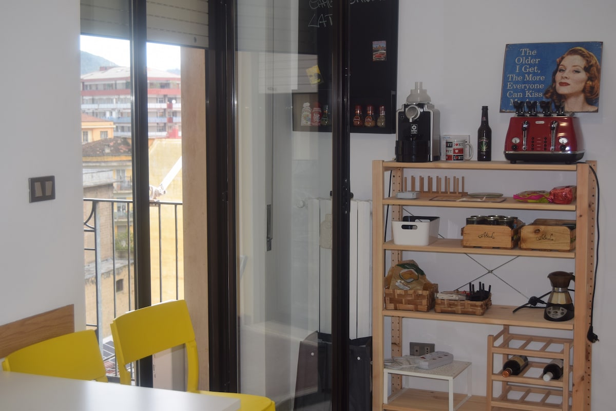 A bright dining area features a table with yellow chairs and a shelving unit displaying cookware and bottles. Large sliding glass doors provide access to a balcony, allowing natural light to fill the space. A coffee maker and a red toaster are visible on the counter.