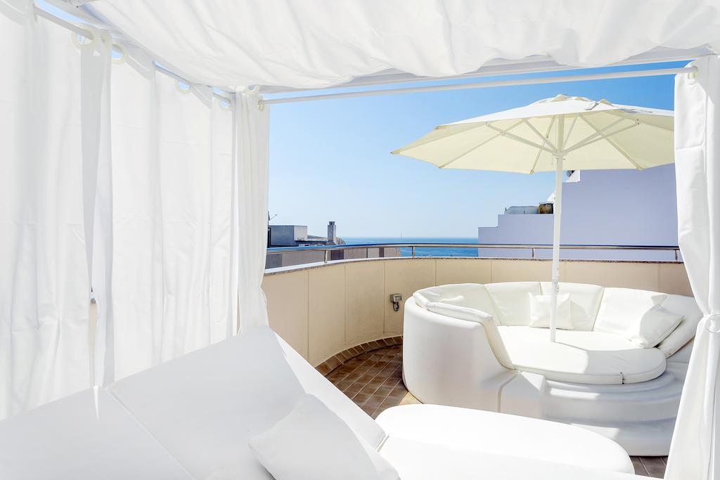A roof terrace is depicted featuring a circular white seating area and a large umbrella offering shade. The expansive view includes the ocean visible in the distance, complemented by a clear blue sky. Light, flowing curtains provide a sense of privacy while allowing natural light.