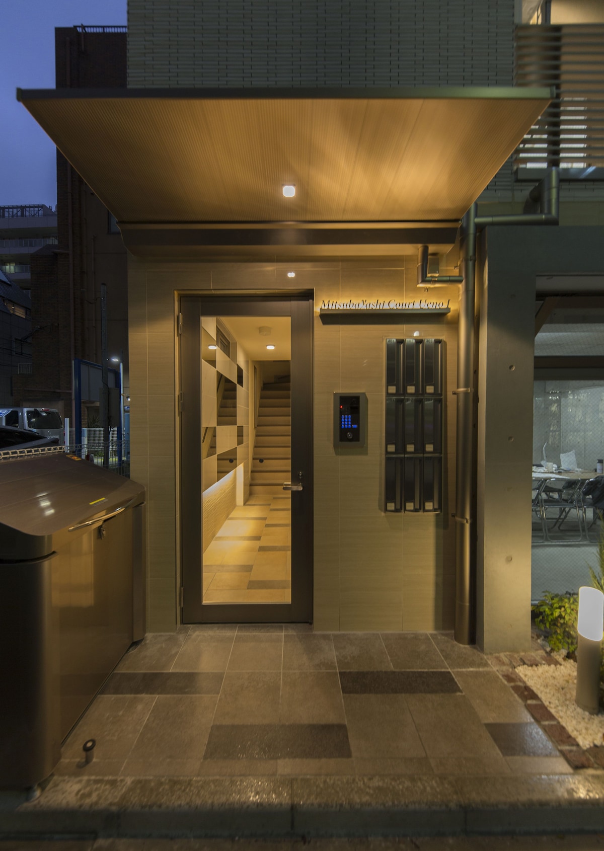 The entrance to the building is illuminated by warm lighting, showcasing a modern door with a keypad entry system. A row of mailboxes is positioned next to the entrance, with a stone pathway leading to the steps inside, providing easy access to the upper floors.