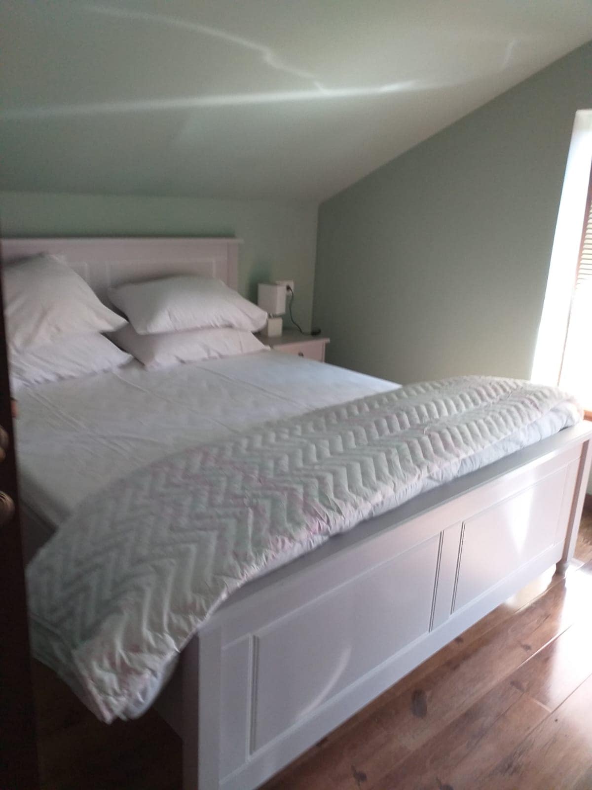 A spacious bed is positioned against a slanted wall, featuring plush pillows and a light comforter. Natural light filters in through a nearby window with wooden blinds, illuminating the soft green wall color and wooden flooring.