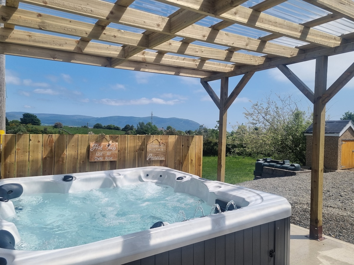 An outdoor hot tub is situated under a wooden pergola, offering an inviting space for relaxation. Scenic views of rolling hills and blue skies are visible in the background, while a wooden fence encloses the area, enhancing privacy.