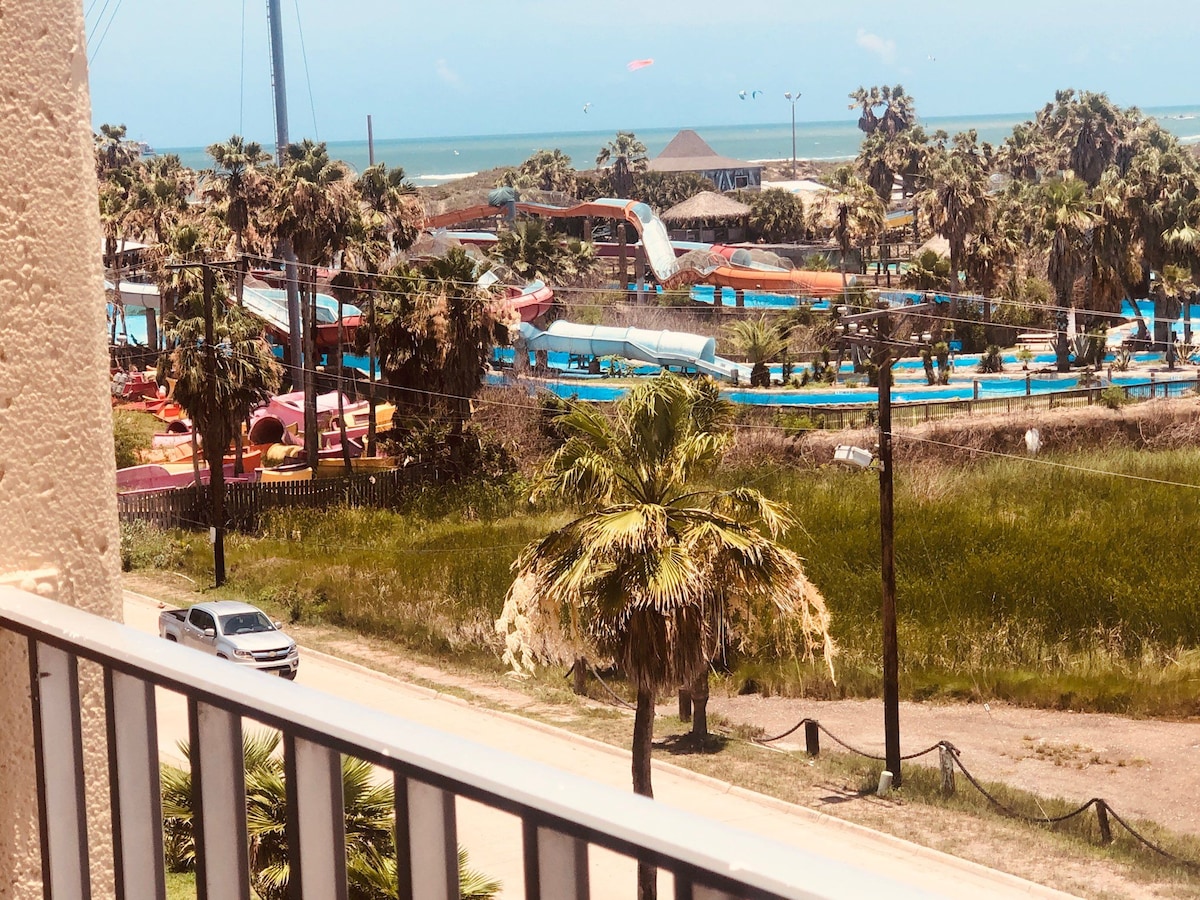 A view from the balcony reveals a colorful water park, surrounded by landscaped palm trees and greenery. The ocean is visible in the background, with a clear blue sky. A road is visible in the foreground, leading to the water park.