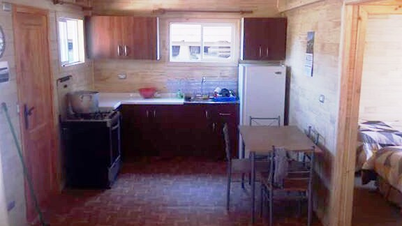 A functional kitchen area is visible, featuring wooden cabinets and a white countertop. A sink, refrigerator, and stove are included. A simple dining table with four chairs is positioned nearby, with light streaming in through two windows that allow for natural illumination.