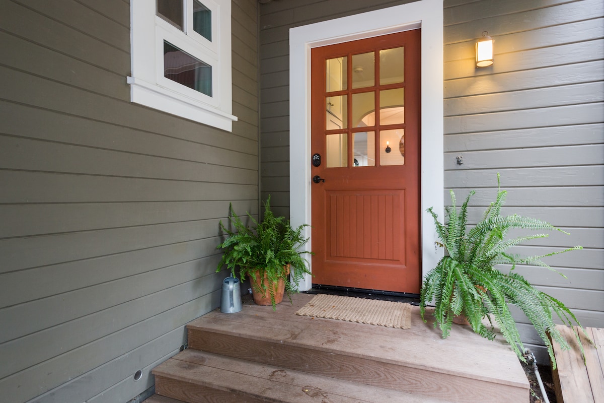 A welcoming entrance is highlighted by a vibrant orange door with multiple small windows, framed by two potted ferns on either side. Wooden steps lead up to the door, while a natural mat sits at the base, accentuating the inviting feel of the entryway.
