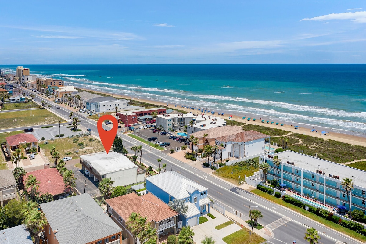 An aerial view captures the proximity of a beach condo to the ocean. The shoreline and colorful beach umbrellas are visible, as well as nearby buildings and streets. The clear blue sky and vibrant waters contribute to a coastal atmosphere.