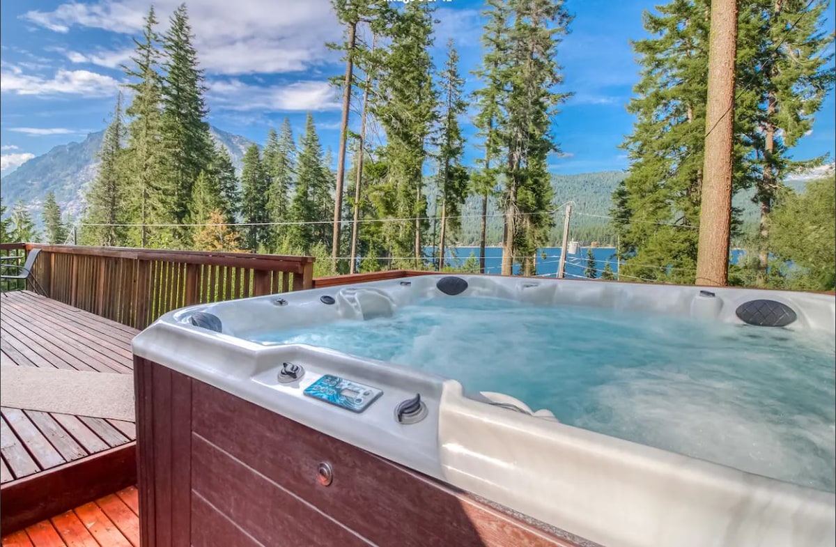 A hot tub is set on a wooden deck, offering a serene view of a forested landscape and Lake Wenatchee in the distance. Towering evergreen trees frame the scene, and the clear blue sky enhances the peaceful atmosphere.