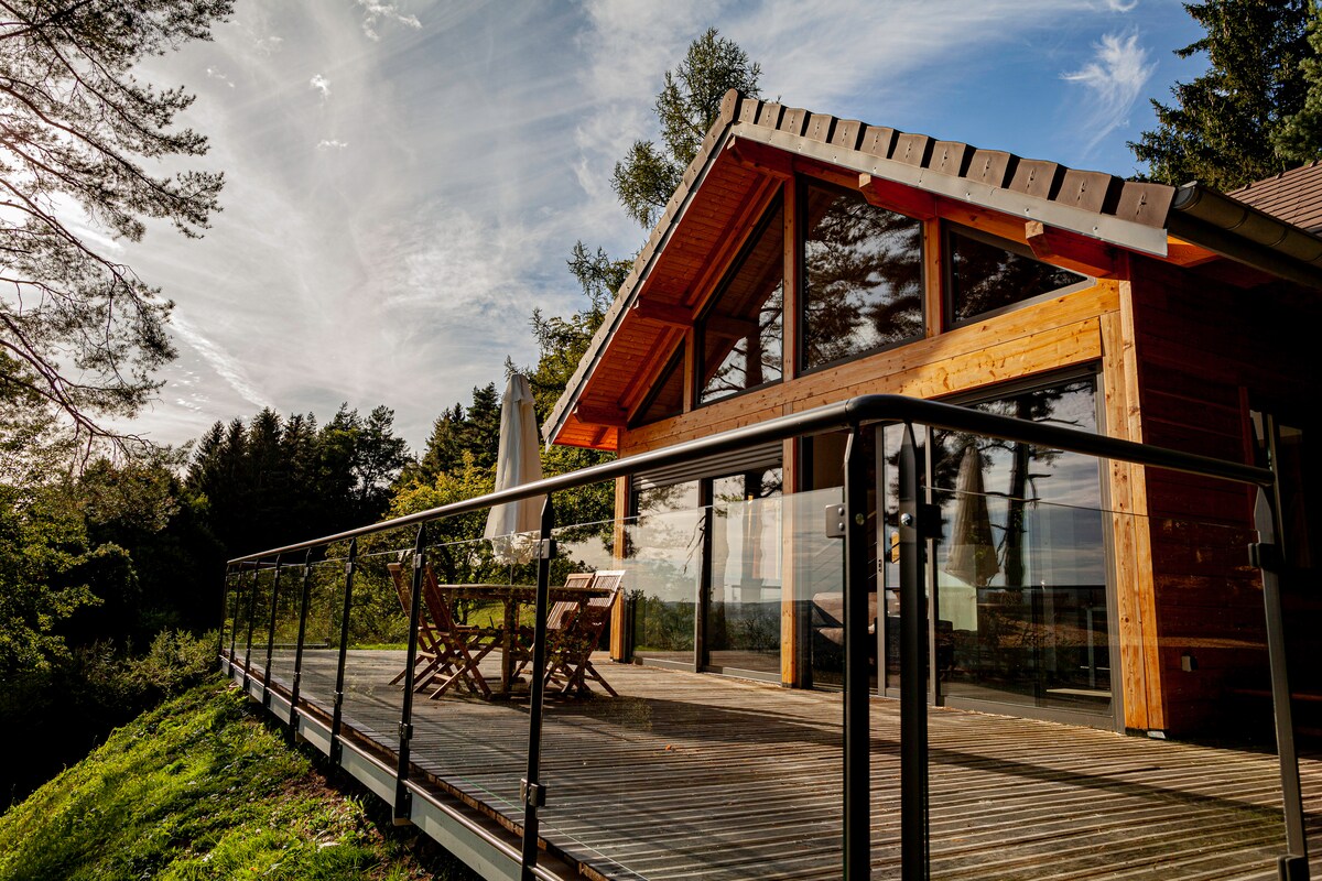A wooden chalet features a spacious deck with a glass railing, showcasing a dining set made of natural wood. Large floor-to-ceiling windows invite ample natural light and provide scenic views of the surrounding trees and landscape.