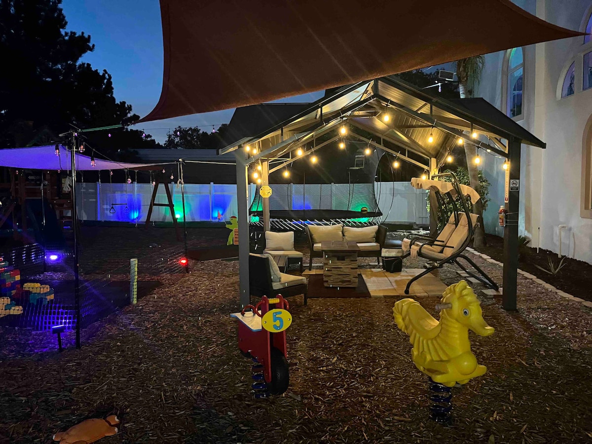 A shaded outdoor seating area is illuminated by string lights, featuring comfortable seating options. Nearby, a colorful playground with a yellow rocking horse and various play structures is visible. The space is designed for relaxation and entertainment, surrounded by a tranquil atmosphere.