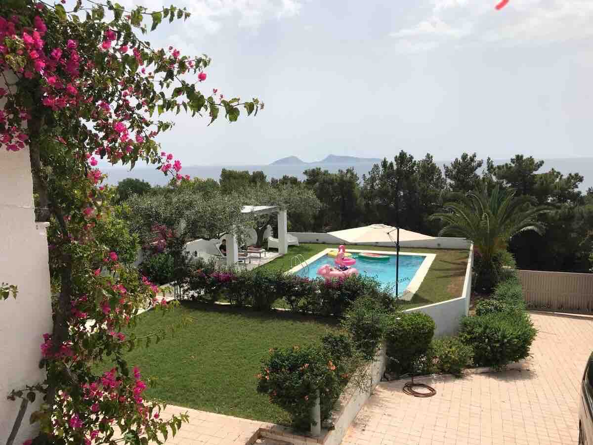 A well-maintained garden showcases a vibrant mix of flowers and green shrubs, with a private pool positioned at the center. Two pink floaties rest on the water's surface, while distant islands and lush trees frame the serene view.