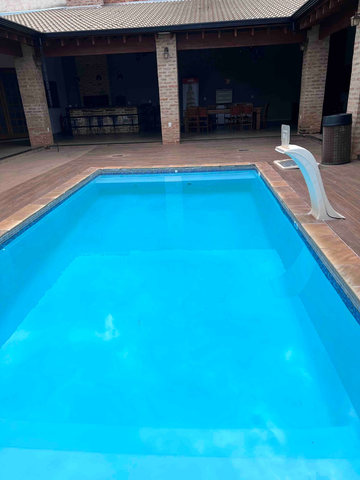 A clean swimming pool is surrounded by a wooden deck, with the water reflecting a bright blue hue. A diving board is positioned at one end, and a shaded area is visible in the background, featuring seating arrangements and dining space.
