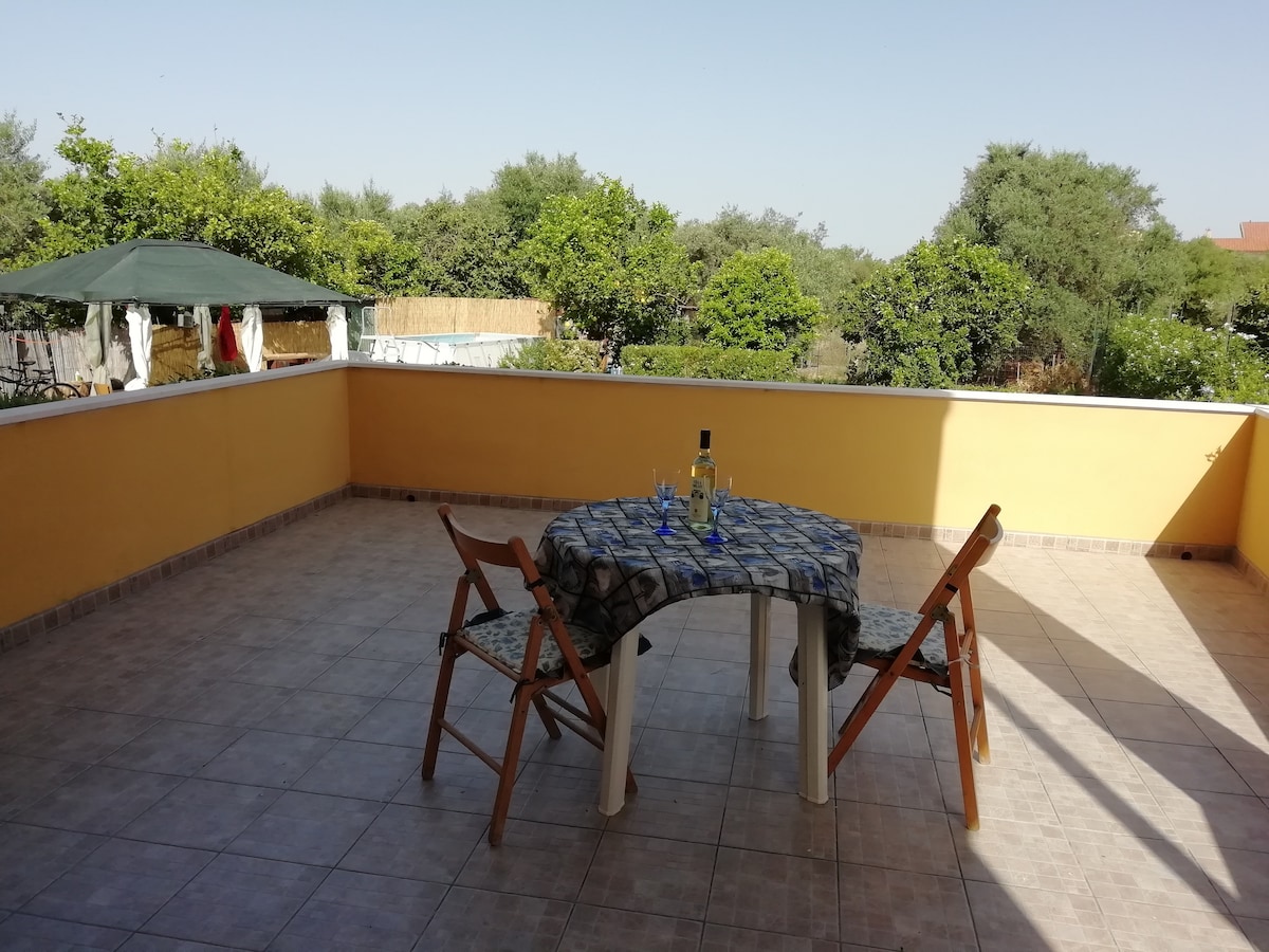 An outdoor terrace is presented, featuring a round table set with two chairs and a bottle of wine. Green vegetation and trees provide a natural backdrop, while the unobstructed sky enhances the sense of spaciousness. The tiled floor extends across the terrace.