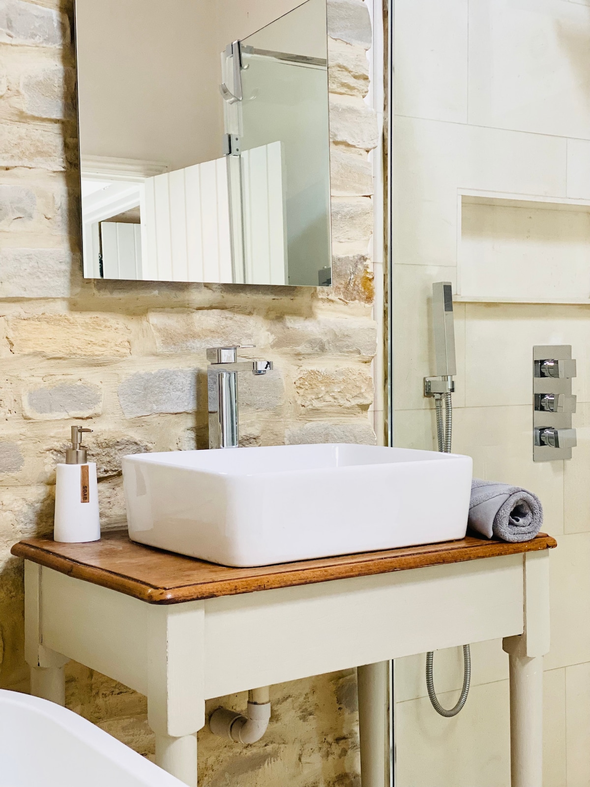 A stylish bathroom features a wall-mounted mirror above a rectangular basin atop a wooden vanity. The space is complemented by stone walls and a walk-in shower with sleek fixtures. A neatly rolled towel is placed beside the basin for convenience.