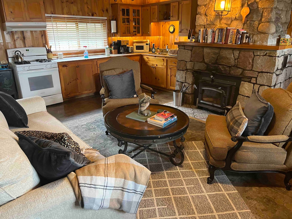 A comfortable living area features a sofa and two armchairs arranged around a circular coffee table, which holds books and a decorative item. A stone fireplace stands against the wall, and the kitchen area is visible in the background, showcasing wooden cabinetry and modern appliances.