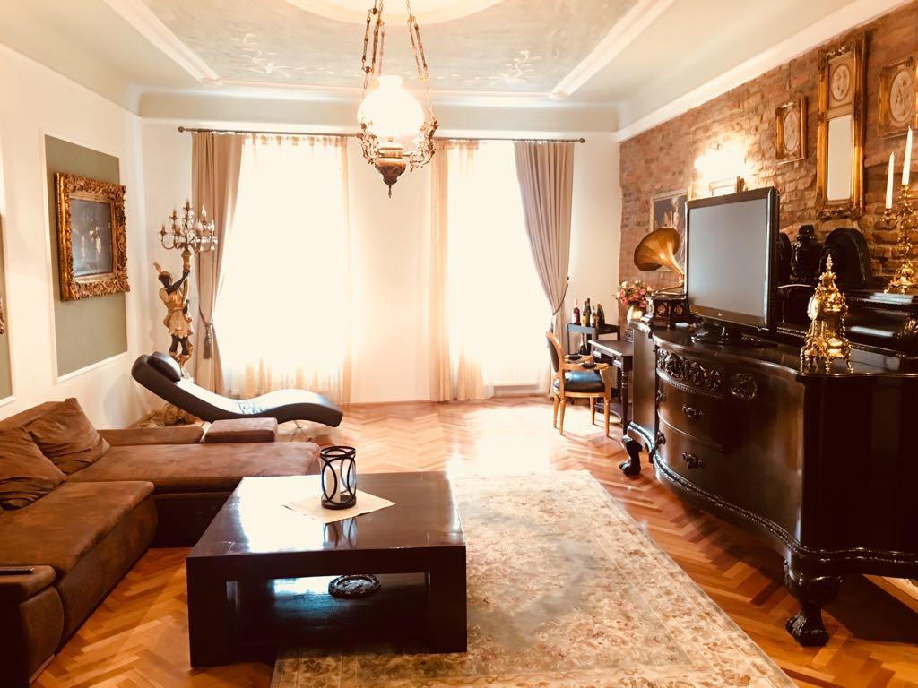 A spacious living area features a modern sofa, a decorative coffee table, and a vintage-style cabinet. Natural light enters through large windows, illuminating the room's rich wooden flooring. Elegant wall art and antique decor add character to the inviting space.