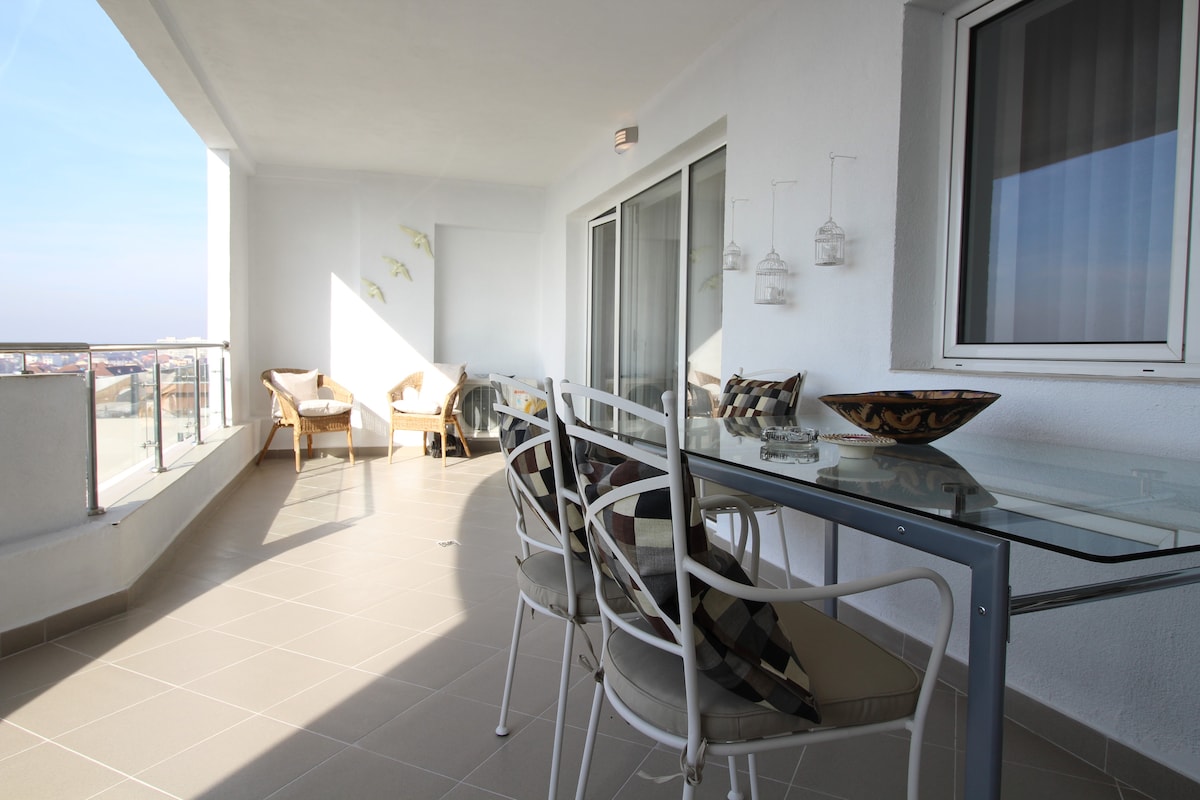 A spacious terrace is presented with a glass table surrounded by comfortable chairs. Sunlight fills the area, while decorative lanterns hang from the wall. The view extends beyond the railing, showcasing the surrounding landscape of Bucharest.