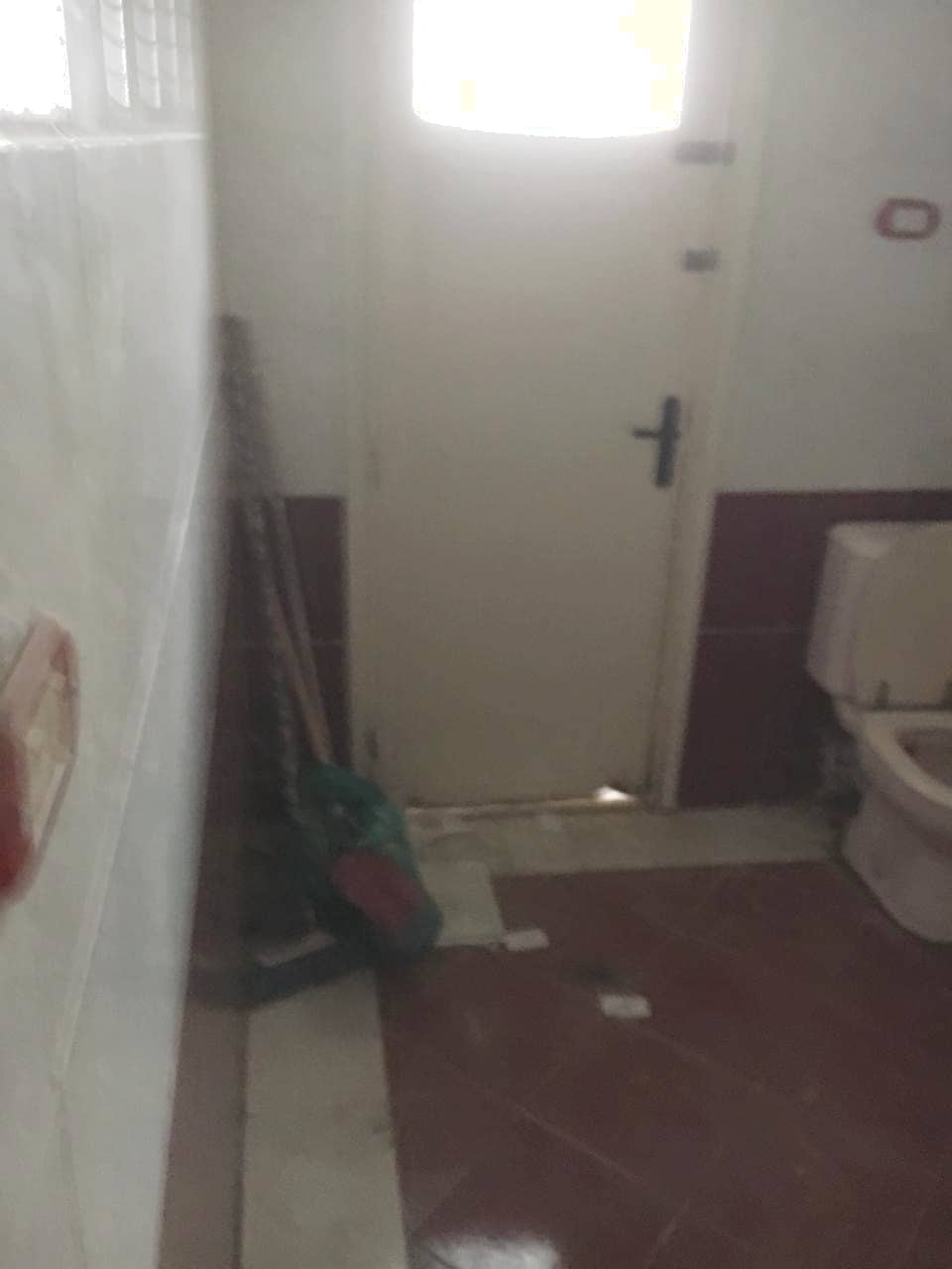 A bathroom is visible featuring a white toilet and a door partially open, allowing light to enter. A cleaning tool is propped against the wall beside the toilet. The floor is finished with ceramic tiles, combining light and dark colors.