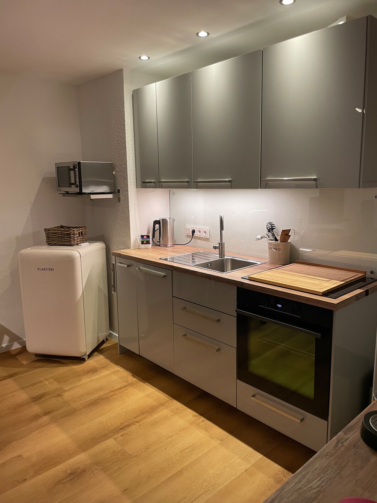 A modern kitchen is equipped with sleek gray cabinetry, a stainless steel sink, and an oven. A wooden cutting board sits atop the counter, alongside a kettle and kitchen utensils. A white refrigerator is positioned to the side, providing ample storage space.