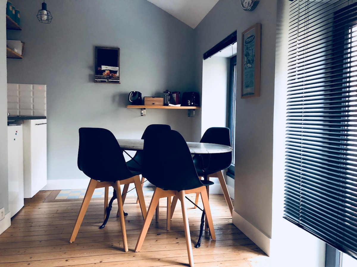 A dining area is visible, featuring a round table surrounded by four black chairs. A small shelving unit displays appliances, while a window allows natural light to enter, complemented by light-colored wooden flooring. Soft gray walls create a calming atmosphere.