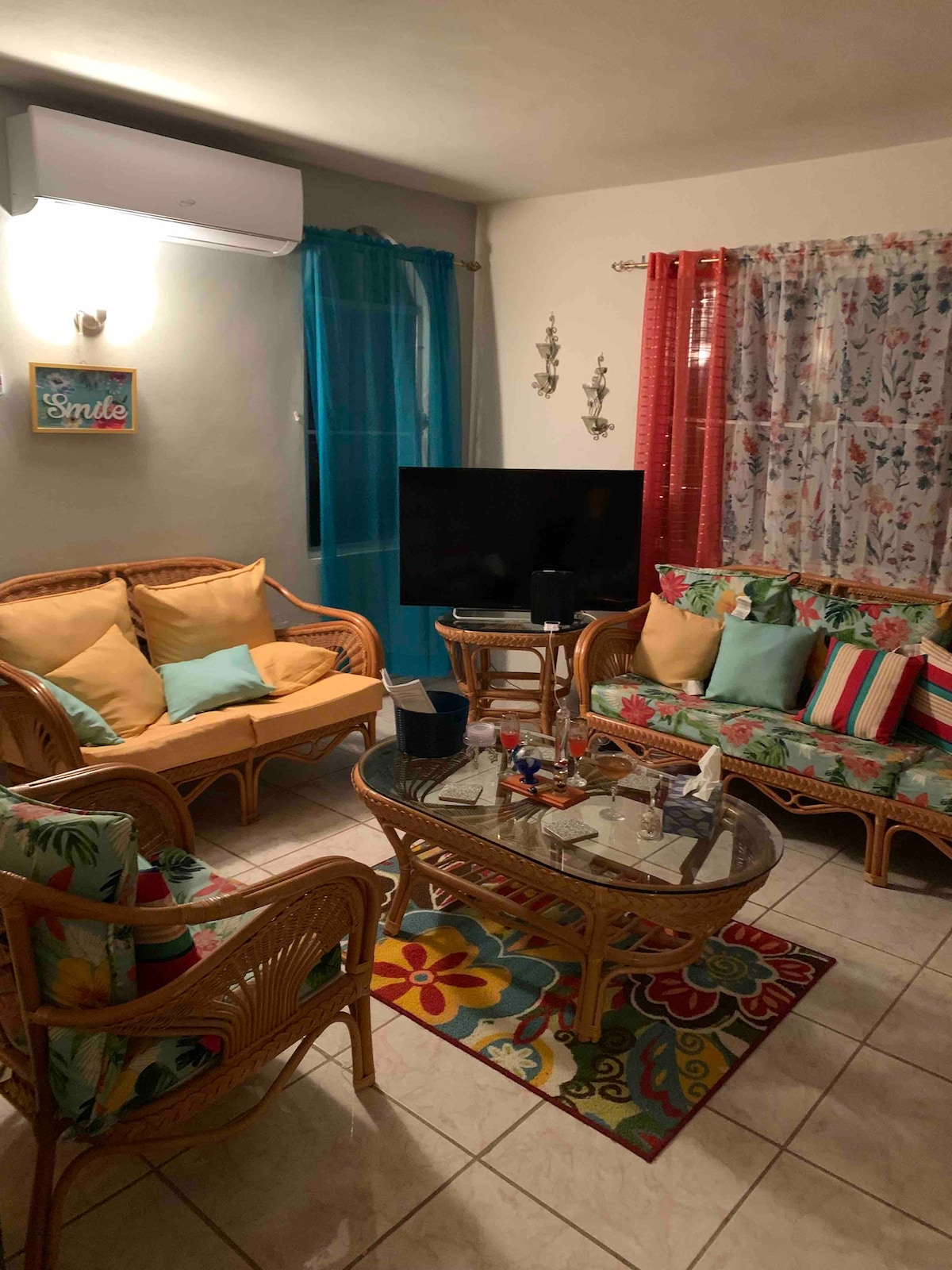 A cozy living area features rattan furniture with colorful cushions, complemented by a glass coffee table. A television is positioned against a neutral wall, while playful patterned curtains add a vibrant touch. Light fixtures create a warm ambiance, enhancing the inviting space.
