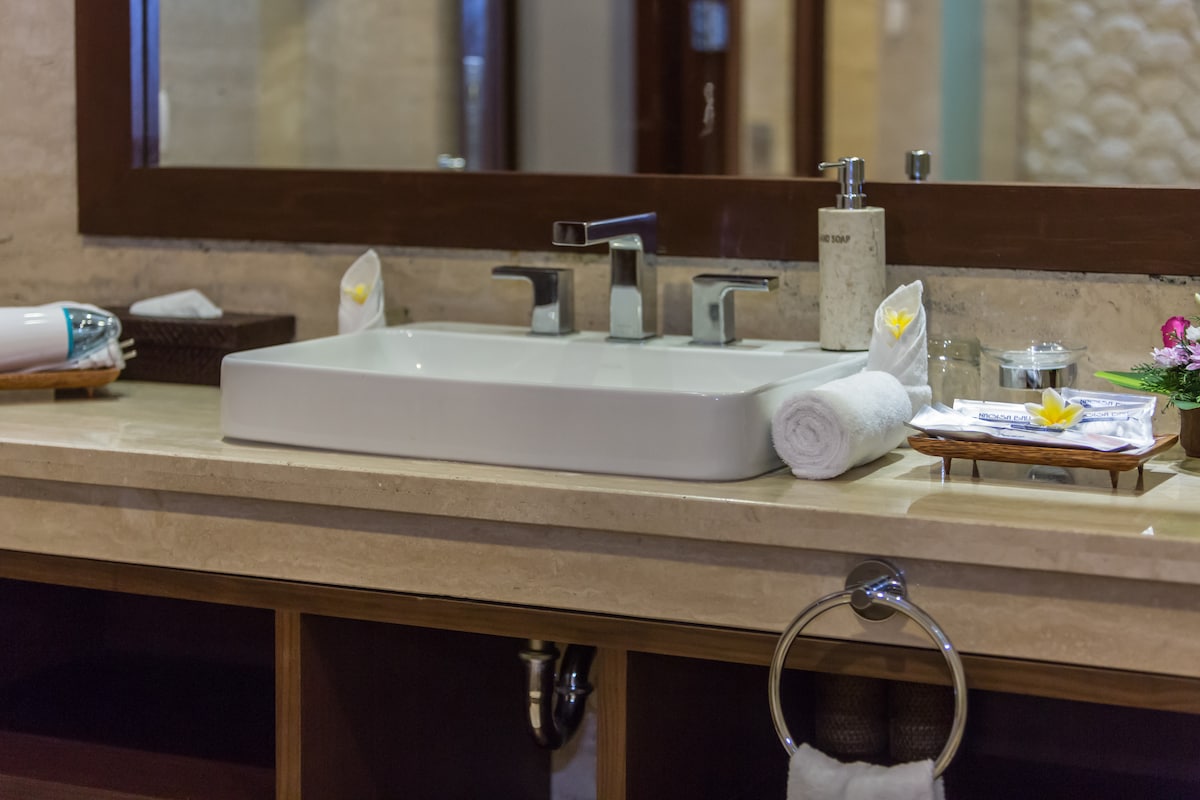 A modern bathroom sink is set atop a marble countertop, featuring minimalist fixtures in polished chrome. Fresh towels are neatly rolled beside a small tray, which holds complimentary toiletries and a decorative flower. A large mirror reflects the elegant ambiance of the space.