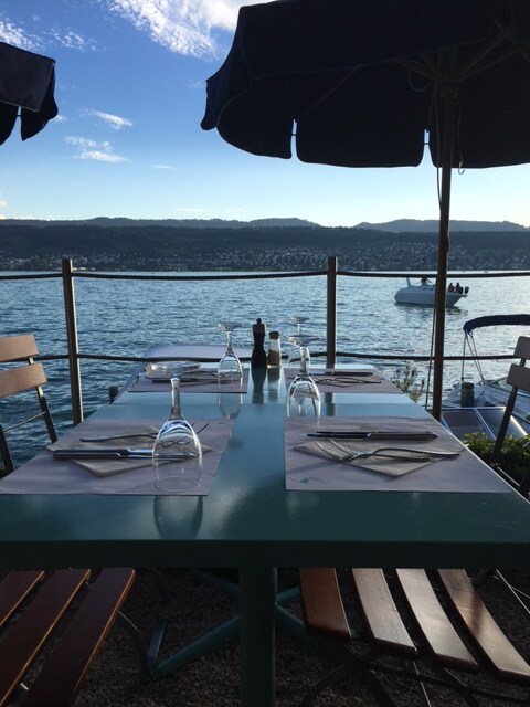 A dining table set for four is positioned on a terrace overlooking the water. Sun umbrellas provide shade, while glassware and utensils are neatly arranged. A scenic view of the lake and distant hills enhances the serene dining atmosphere.