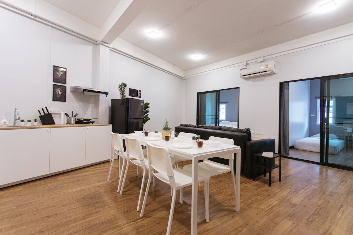 A spacious kitchen and dining area features a white table set for six, surrounded by modern chairs. A black sofa is visible in the living space, with kitchen appliances and cabinets neatly arranged. Large glass doors lead to the bedrooms, enhancing natural light.