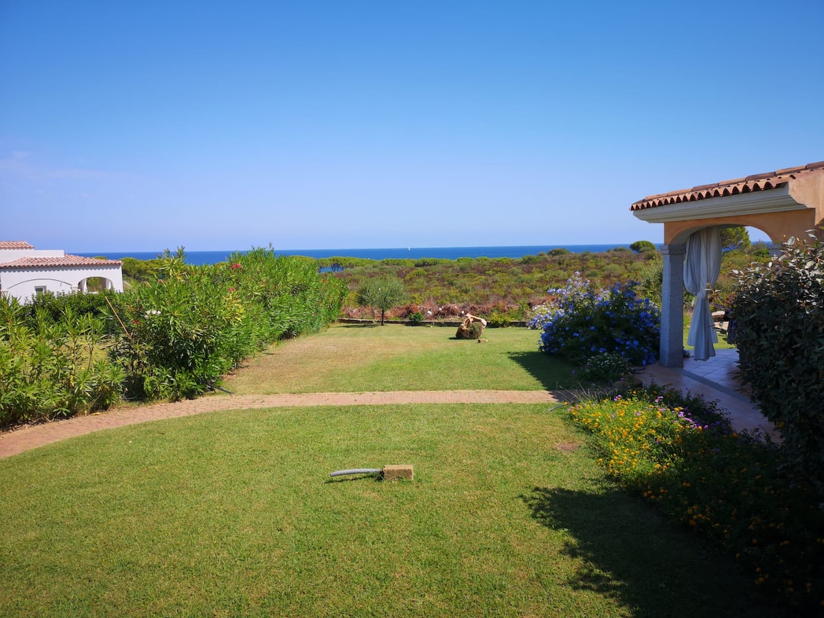 A spacious garden filled with vibrant greenery and various flowers extends toward the sea. A covered veranda is visible on the right, providing a shaded area to enjoy the seaside view. The horizon blends softly with the blue sky, enhancing the serene atmosphere.