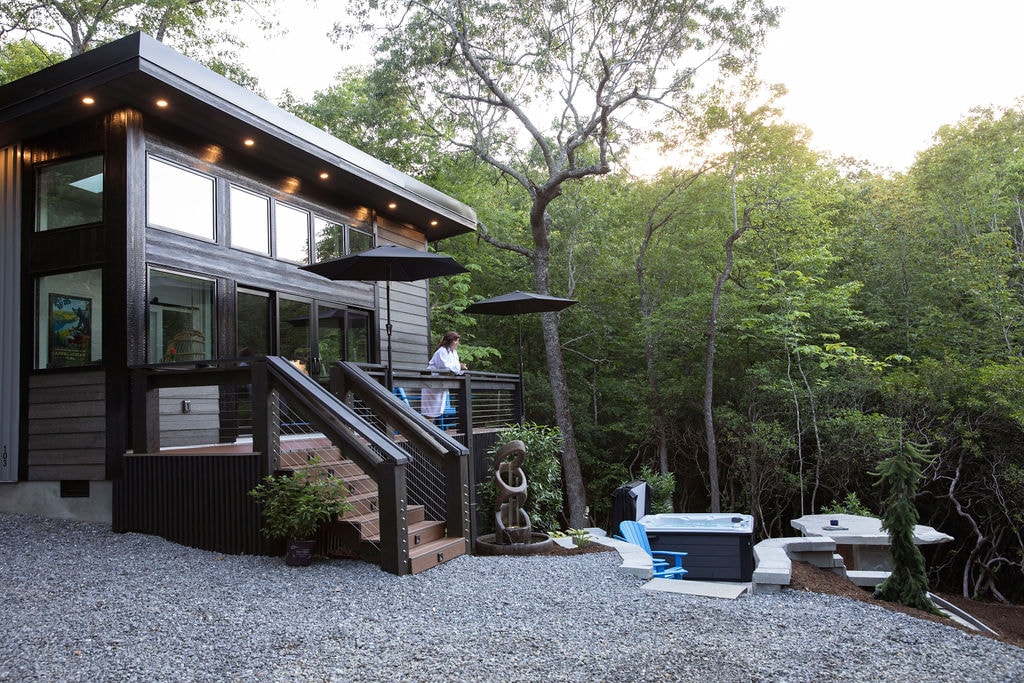 The contemporary mountain home is set against a backdrop of lush trees, featuring a gravel pathway leading to a wraparound deck. Two outdoor seating areas are visible, along with a private hot-tub nestled nearby, providing a peaceful retreat.