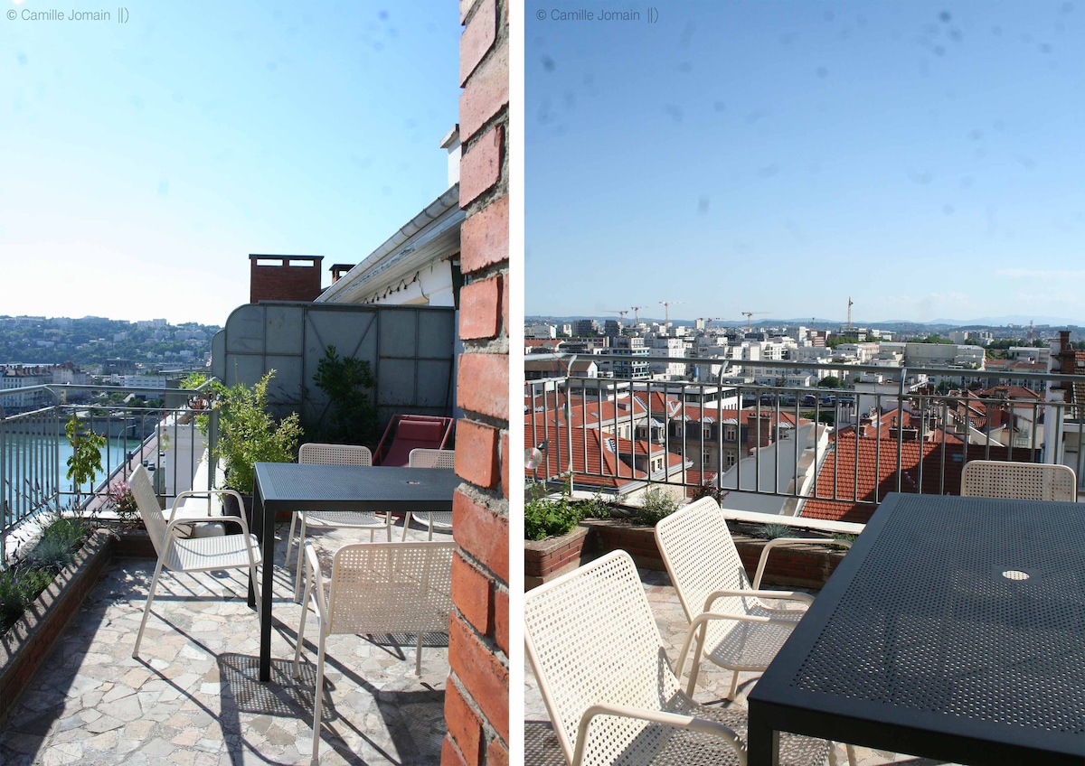 A spacious terrace showcases outdoor furniture, including a table and chairs. Surrounding views of the city skyline, rooftops, and distant hills are visible against a clear blue sky, providing an inviting space for relaxation or dining.
