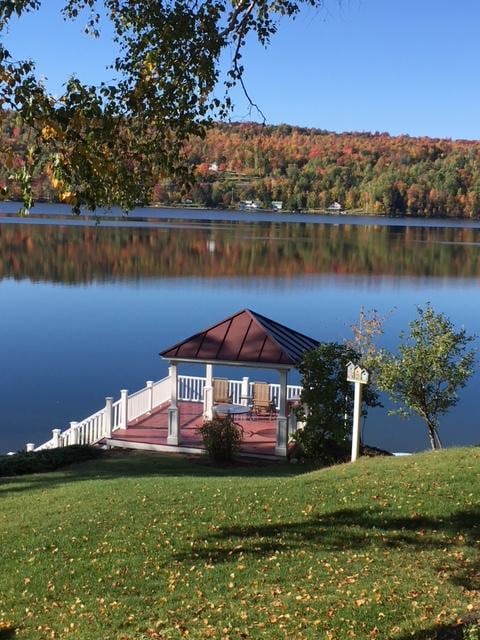 A serene lakeside gazebo is showcased, featuring a raised platform with a peaked roof and surrounding railings. Two comfortable chairs are positioned invitingly, while the calm water reflects the vibrant autumn foliage in the background. Lush green grass complements the scenic view.
