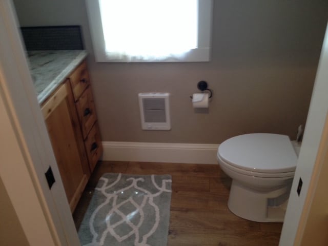 A bathroom space is shown featuring natural wood cabinetry and a countertop with a marbled pattern. A white toilet sits adjacent to a plush area rug. A window with soft fabric covers allows natural light to enter, enhancing the room's airy feel.