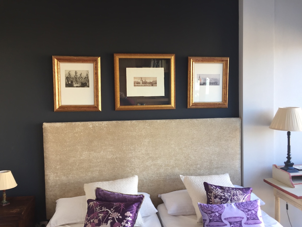 A cozy sleeping area features two spacious beds topped with decorative pillows in a soft purple hue. Above the beds, three framed artworks adorn the dark accent wall, complemented by a bedside lamp casting a gentle glow and stacks of books nearby.