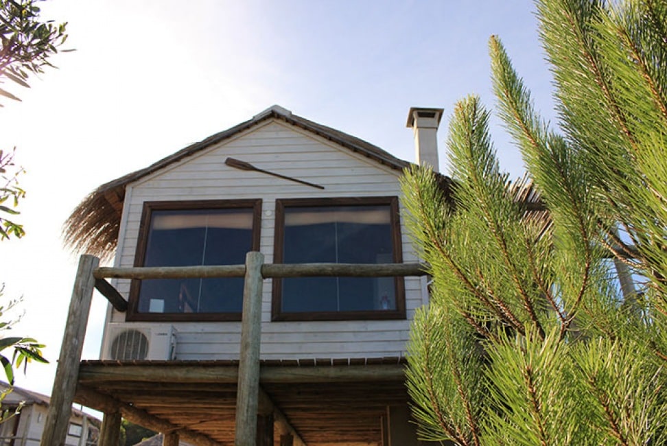 A charming cabin is set on stilts, featuring large windows that provide ample natural light. The exterior is accented by wooden beams and a decorative paddle. Lush greenery surrounds the cabin, adding to the secluded atmosphere and inviting a connection with nature.