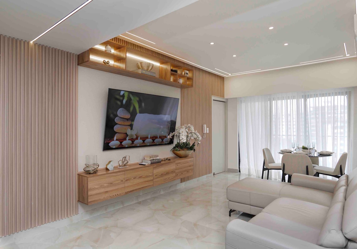 A modern living area features a comfortable sofa and a large wall-mounted TV above a wooden media console. Natural light filters through sheer curtains, illuminating the space. A dining table with four chairs is visible in the background, enhancing the open layout.