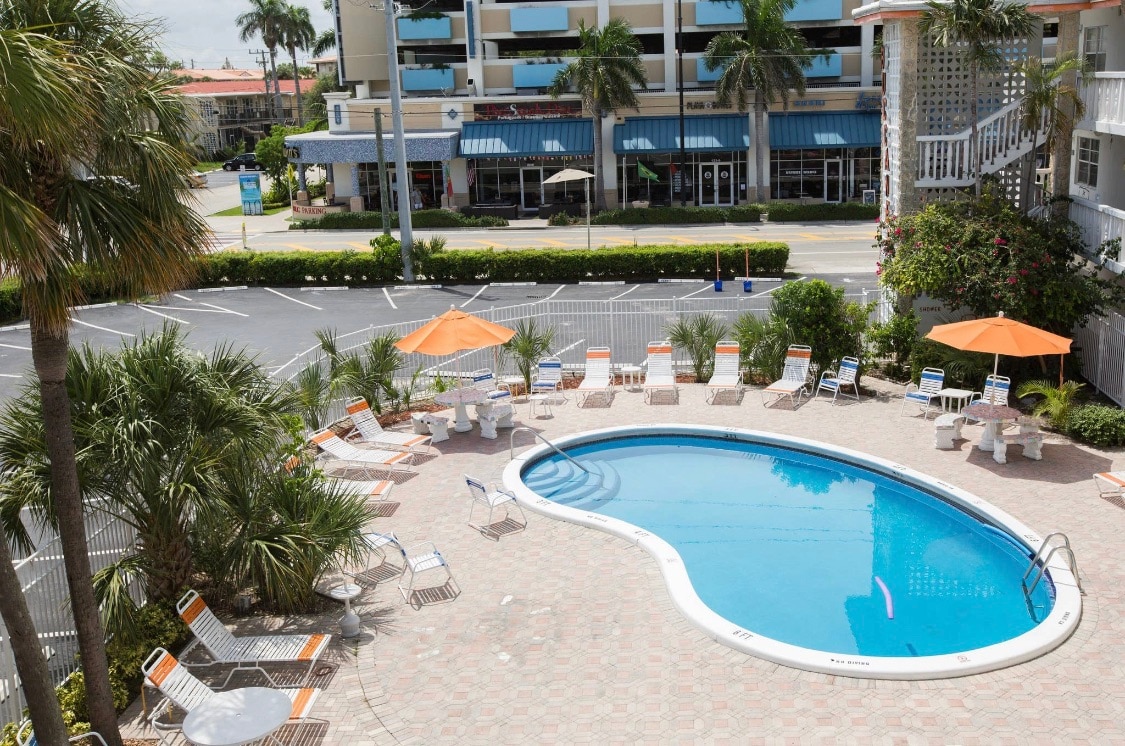 A circular pool is surrounded by lounge chairs and umbrellas, creating a relaxing outdoor space. Palm trees add a tropical touch, while nearby buildings are visible in the background. The area is thoughtfully designed for leisure and social gatherings.