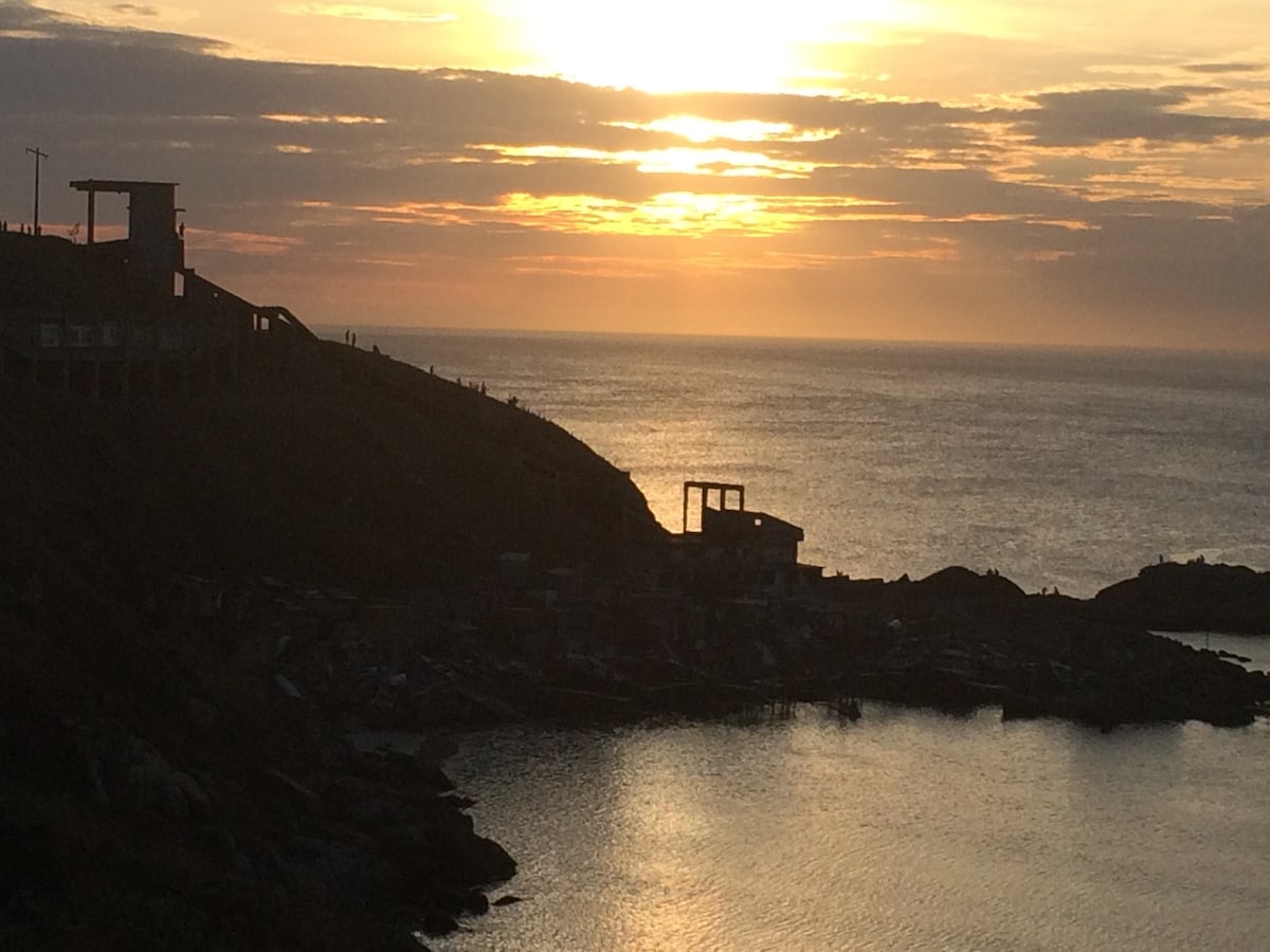 A scenic coastal view captures the sun setting over the horizon, reflecting golden hues on the water's surface. Silhouettes of distant hills and structures are visible, while gentle waves lap at the shore, creating a serene atmosphere.
