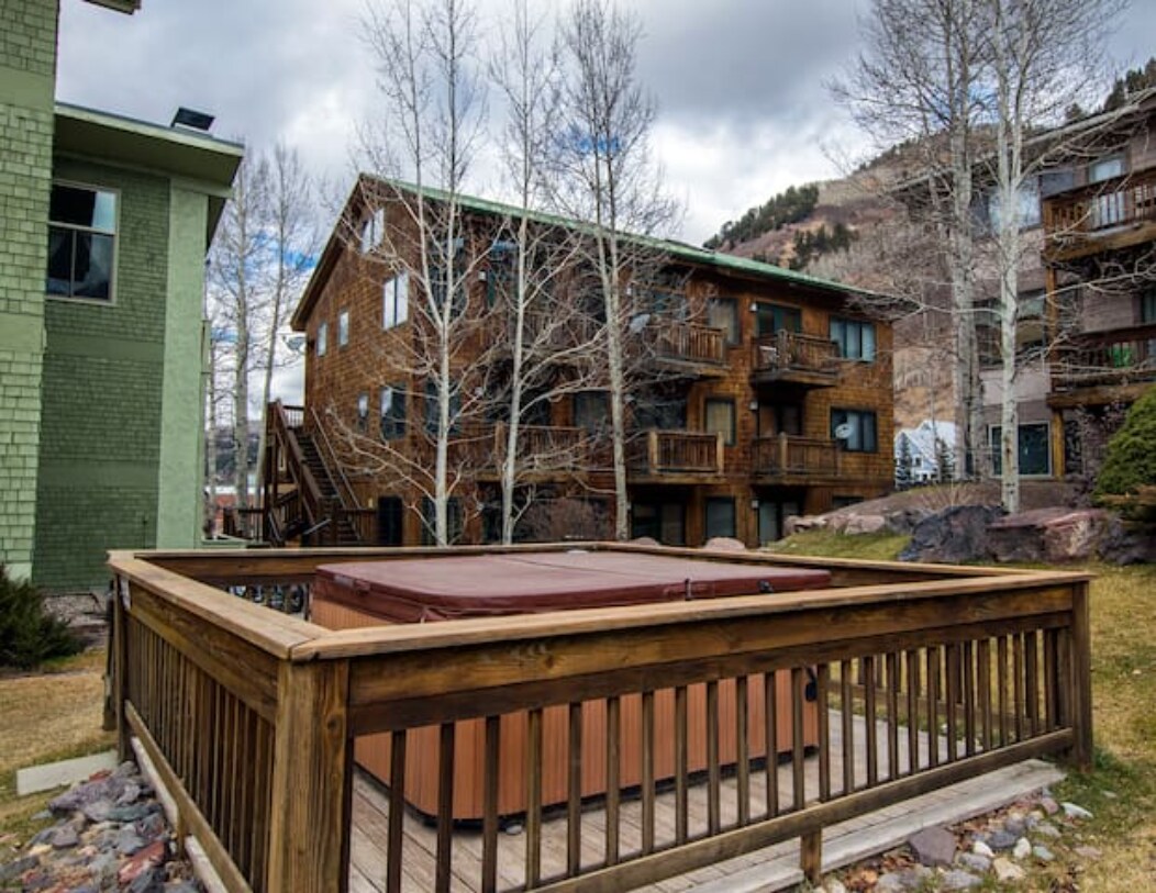 An outdoor hot tub is enclosed by wooden decking, offering a space for relaxation amidst tall Aspen trees. Surrounding buildings are visible, showcasing a variety of architectural styles and natural elements in the backdrop of a mountainous landscape.