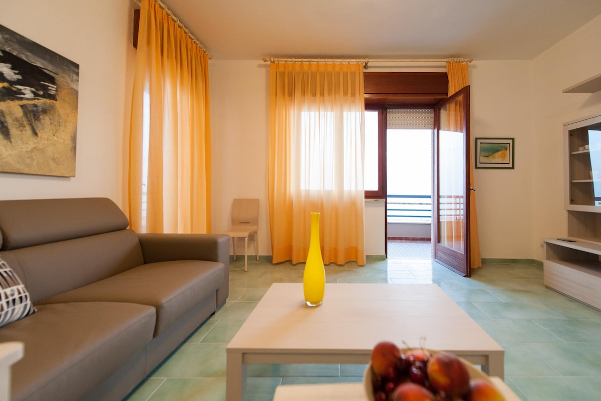 A spacious living area features a grey sofa and a light-colored coffee table. Bright yellow curtains frame large windows that allow natural light to fill the room. A bowl of fresh fruit sits on the table, providing a touch of color to the neutral palette.