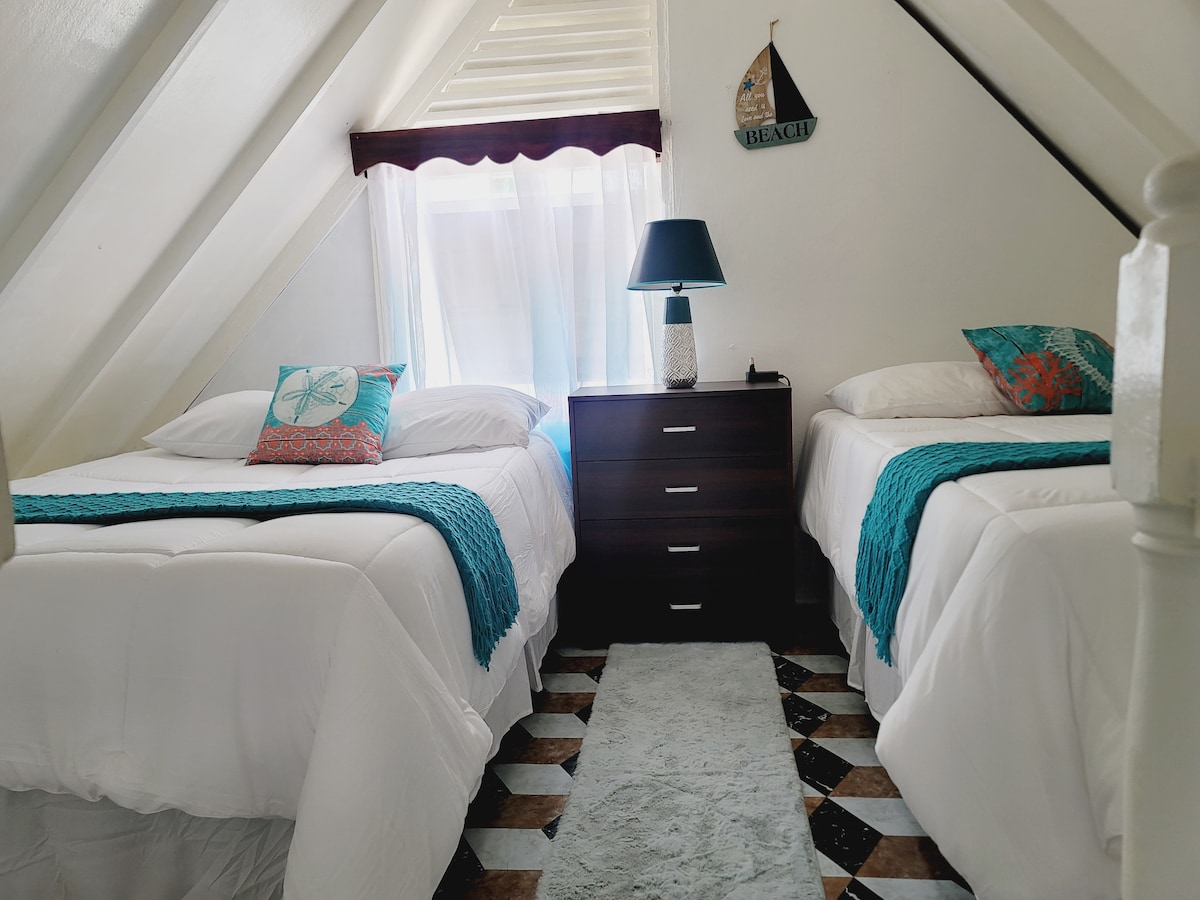 A cozy sleeping area features two spacious beds adorned with decorative pillows and soft blankets. A small chest of drawers is positioned nearby, complemented by a lamp and natural light filtering through the bright window with white curtains.