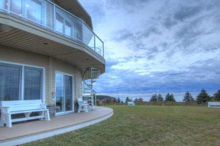 The exterior of the modern condo is captured, featuring a spiral staircase ascending to the upper level. A spacious deck is bordered by a well-maintained lawn, with white benches positioned for seating. The sky displays a mix of clouds, creating a serene backdrop.