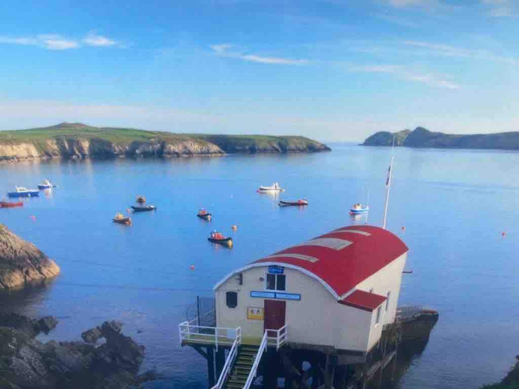 A tranquil seaside scene features a red-roofed boat house situated on the water's edge. Several boats are anchored in the calm bay, surrounded by rocky shores and distant hills under a clear blue sky.