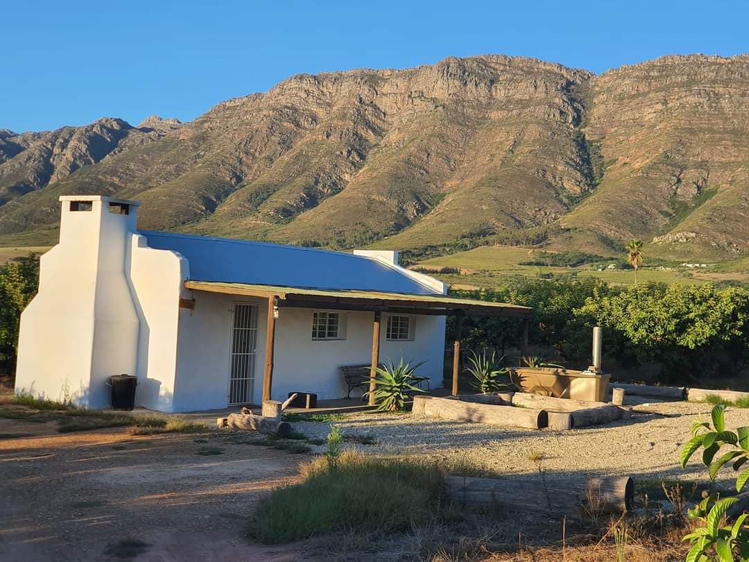 A charming cottage is set against a backdrop of towering mountains, surrounded by lush greenery. A blue roof contrasts with the white walls, while large windows offer views of the outdoor landscape. An outdoor seating area is positioned nearby, inviting relaxation.